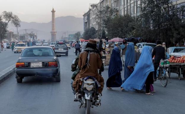 Un grupo de mujeres vestidas con burka cruzan una carretera en Kabul ante dos talibanes en moto y armados. 